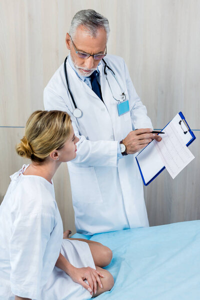 serious male doctor with stethoscope over neck pointing at clipboard to female patient in hospital room 