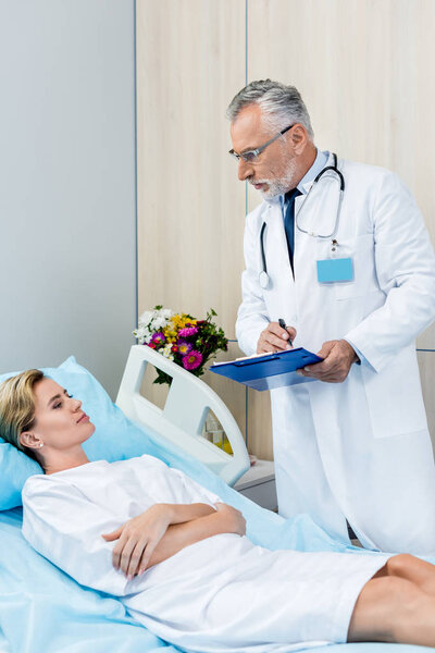 serious male doctor with stethoscope over neck writing in clipboard near adult female patient in hospital room 