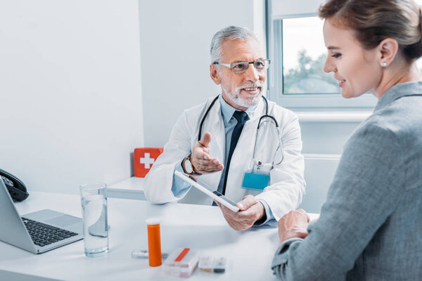 smiling middle aged male doctor with digital tablet gesturing by hand and talking to female patient at table in office 