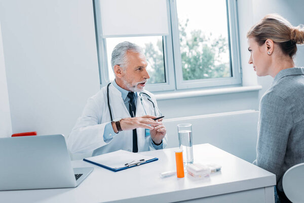 mature male doctor talking to female patient at table with laptop in office 