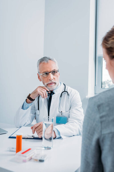 serious mature male doctor talking to female patient at table with pills and clipboard in office 