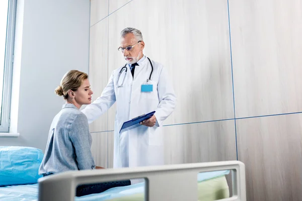 成熟男性医生与剪贴板欢呼女性病人在医院室 — 图库照片