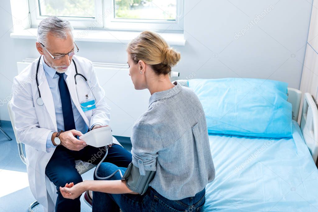 high angle view of mature male doctor with stethoscope measuring pressure of female patient by tonometer in hospital room