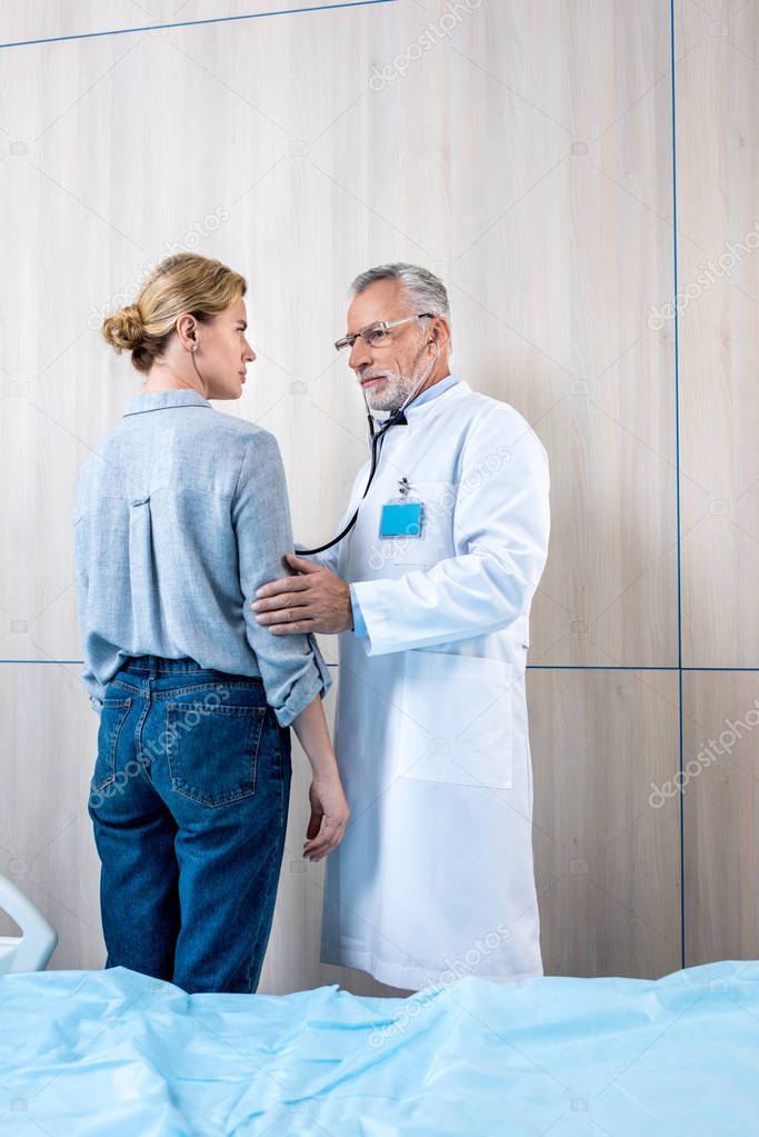 confident mature male doctor examining female patient by stethoscope in hospital room