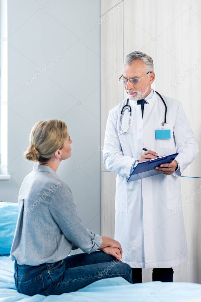 mature male doctor writing in clipboard near female patient in hospital room