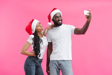 Noel şapkalı kız arkadaşı dilini dışarı izole üzerinde pembe bir arka plan ile smartphone selfie alarak genç Afro-Amerikan adam 