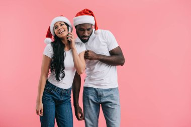 concentrated african american man in chrismas hat eavesdropping girlfriend talking on smartphone isolated on pink background  clipart