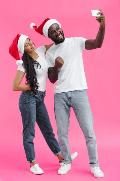年轻的非洲裔美国人男子在圣诞帽显示拳头和采取自拍在智能手机与女朋友在粉红色背景 — 图库照片