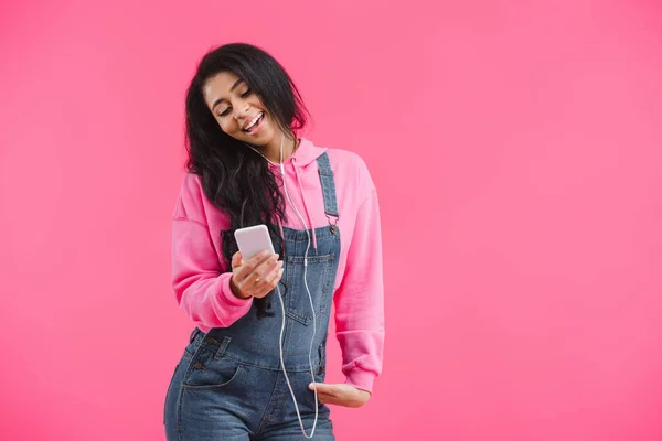 ピンクの背景に分離されたスマート フォンで音楽を聴くイヤホンで幸せな若いアフリカ系アメリカ人女性 — ストック写真