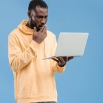 Shocked african american man closing mouth by hand and looking at laptop isolated on blue background