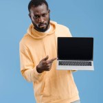 Serio giovane uomo afroamericano che punta il dito al computer portatile con schermo bianco isolato su sfondo blu