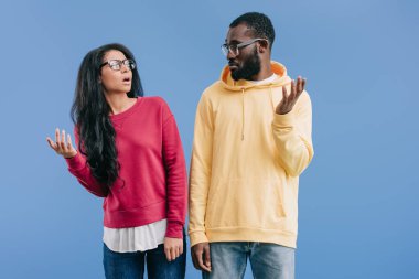 confused african american couple in eyeglasses looking at each other and doing dubium gesture isolated on blue background  clipart