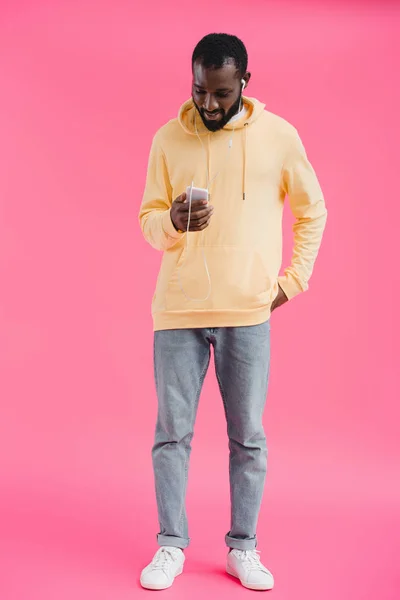 stylish african american man in earphones listening music with smartphone on pink background