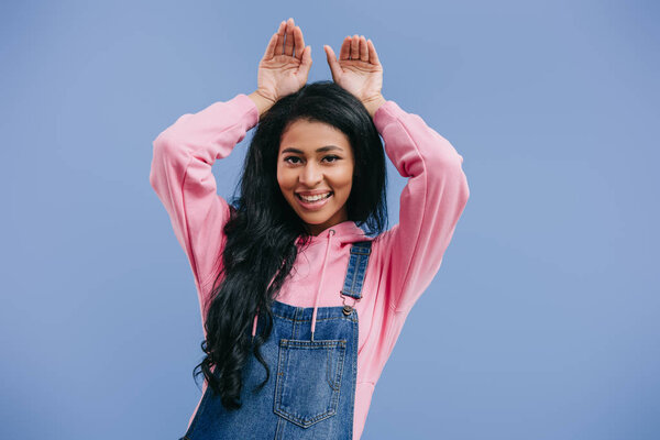 smiling young african american woman doing bunny ears by hands isolated on blue background 