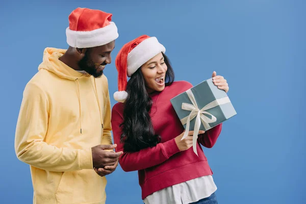 excited african american woman in christmas hat holding gift box while her boyfriend standing near isolated on blue background