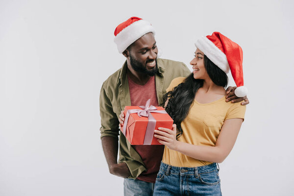 smiling african american couple in santa hats looking at each other and holding present isolated on white