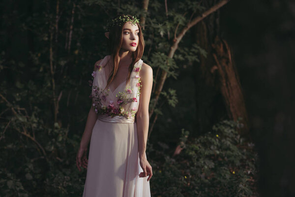 Beautiful mystic elf in flower dress and floral wreath in woods