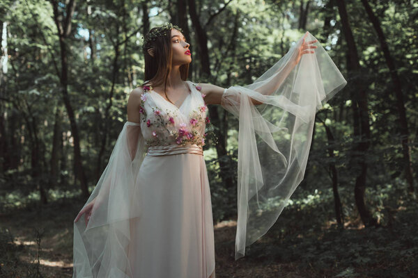 beautiful elf in elegant dress with flowers posing in forest