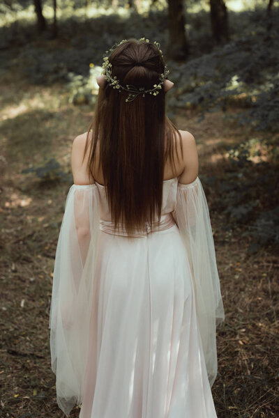 rear view of mystic elf in elegant dress and floral wreath in forest