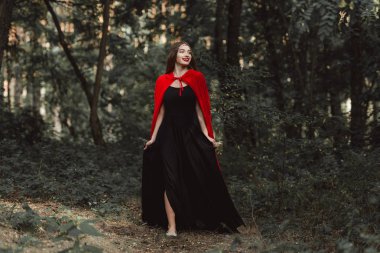 beautiful mystic girl in black dress and red cloak walking in forest clipart