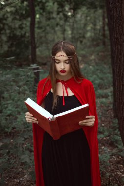 beautiful mystic girl in red cloak and wreath reading magic book in woods clipart