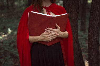 cropped view of mystic girl in red cloak holding magic book in forest clipart