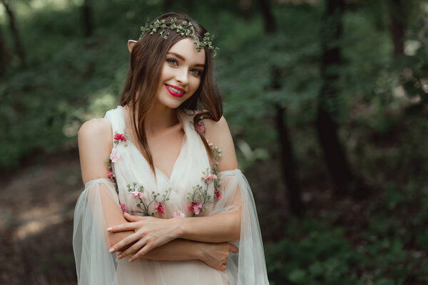 attractive smiling girl in dress and floral wreath posing in woods