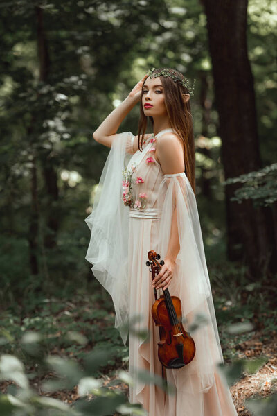 mystic elf in flower dress and floral wreath holding violin in forest