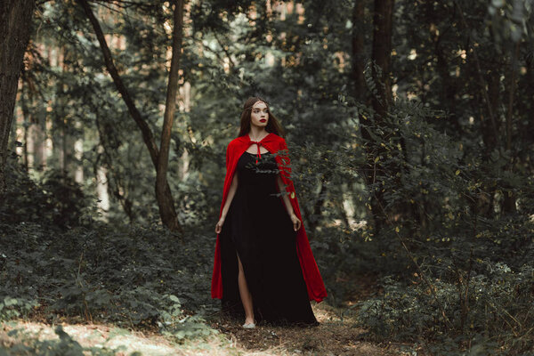 Mystic girl in black dress and red cloak walking in forest