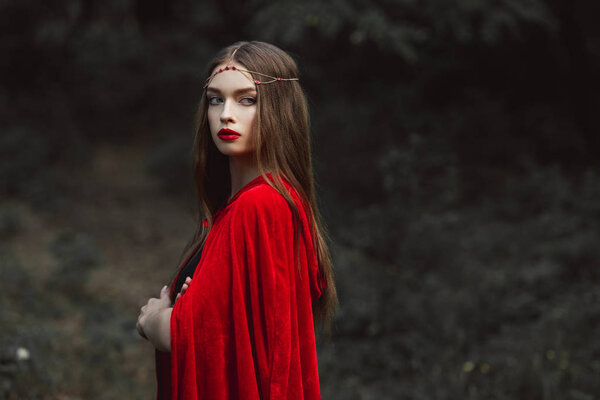 Beautiful girl in red cloak and elegant wreath in woods