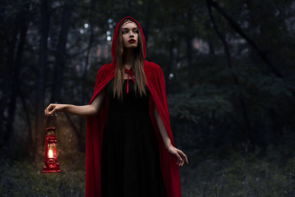 Elegant mystic girl in red cloak with kerosene lamp walking in dark woods