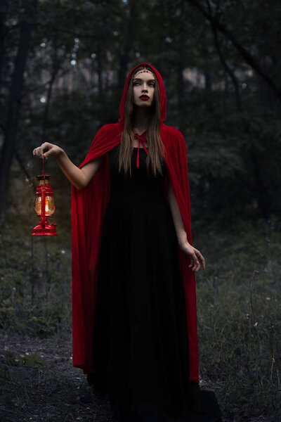 Young mystic woman in red cloak with kerosene lamp walking in dark woods
