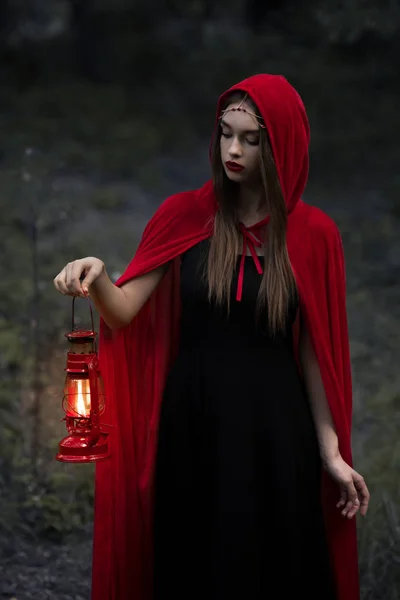 灯油ランプで暗い森を歩く赤いマントで神秘的な美少女 — ストック写真