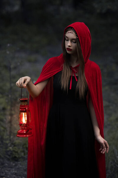 Beautiful mystic girl in red cloak walking in dark forest with kerosene lamp