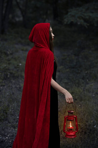 mystic girl in red cloak walking in dark forest with kerosene lamp
