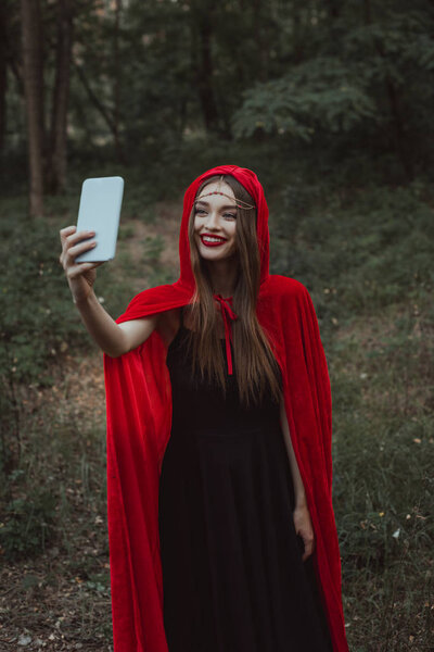 mystic girl in red cloak taking selfie on smartphone in forest