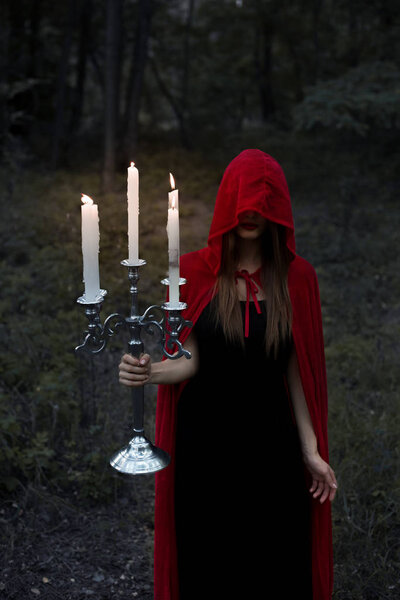 Magic woman in red cloak and hood holding candelabrum with candles in dark forest