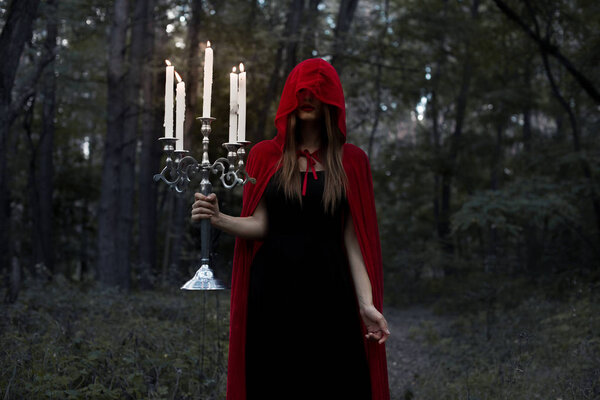 mystic girl in red cloak and hood holding candelabrum with candles in dark forest