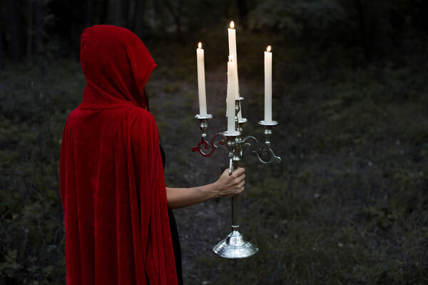 Mystic girl in red cloak holding candelabrum with flaming candles in dark woods
