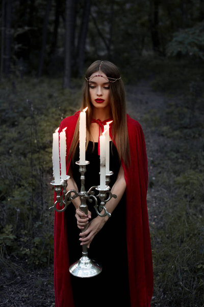 beautiful mystic girl in red cloak holding candelabrum with flaming candles in forest