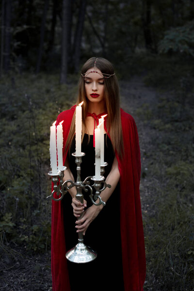 Beautiful mystic girl in red cloak holding candelabrum with flaming candles in forest
