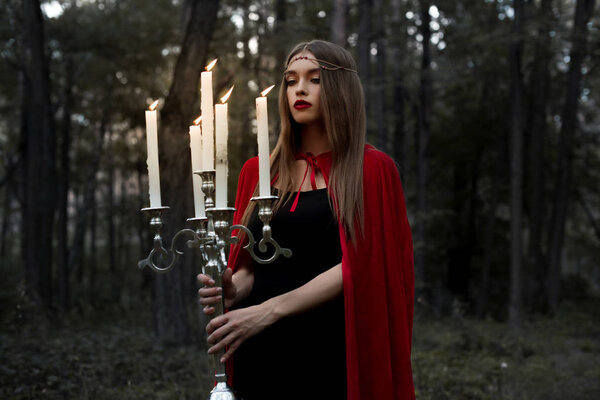 Mystic girl in red cloak holding candelabrum with flaming candles in forest