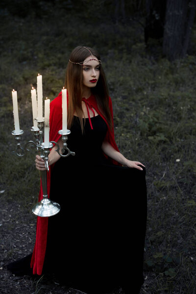 Mystic girl in red cloak holding candelabrum with flaming candles walking in forest