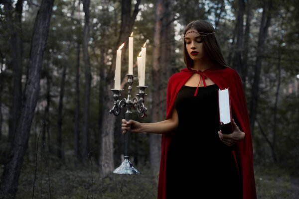 Mystic girl in red cloak holding candelabrum with flaming candles and magic book in dark woods