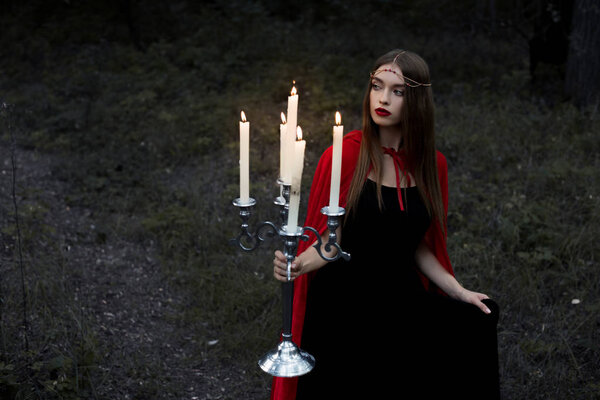 Elegant mystic girl in red cloak holding candelabrum with flaming candles walking in dark forest