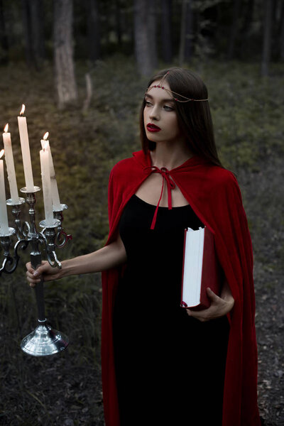 Mystic girl in red cloak holding candelabrum with flaming candles and magic book in forest