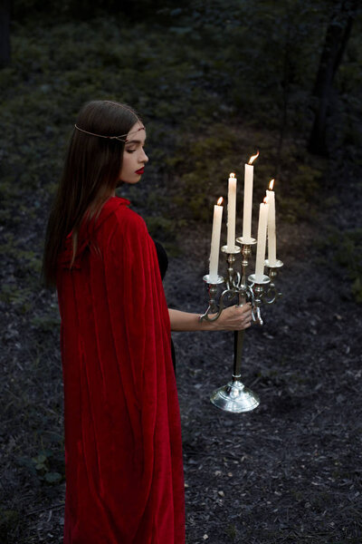 Elegant girl in red cloak holding candelabrum with candles and walking in forest
