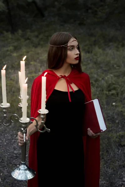 フォレストの炎のキャンドルと魔法の本と燭台を保持している赤いマントで神秘的な美少女  — 無料ストックフォト