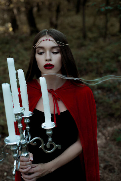 attractive mystic girl in red cloak blowing candles in forest