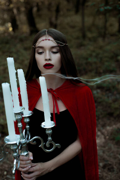 Attractive mystic girl in red cloak blowing candles in forest